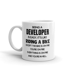 Könnte beinhalten: Eine weiße Keramik-Kaffeetasse mit einem schwarzen Design, auf dem steht: "Ein Entwickler zu sein ist einfach, es ist wie Fahrrad fahren, nur dass das Fahrrad in Flammen steht, du in Flammen stehst, alles in Flammen steht und du in der Hölle bist."