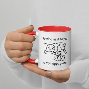 May include: White ceramic mug with a red interior and handle. The mug features a black and white cartoon illustration of two figures in bed, with the text "Rotting next to you is my happy place."