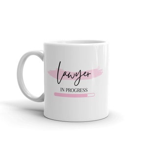 Könnte beinhalten: Weiße Keramiktasse mit einem rosafarbenen Pinselstrich-Design und dem Text "Anwalt in Ausbildung" mit einer rosafarbenen Fortschrittsanzeige.