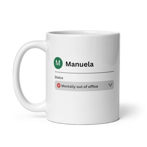May include: White ceramic mug with the name "Manuela" and a green initial "M" printed on the side. The mug also features the status "Mentally out of office" in a gray box.