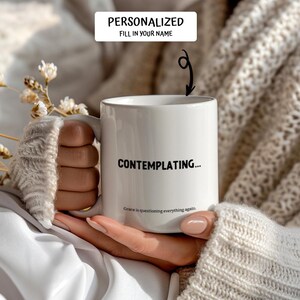 May include: White ceramic mug with the text "CONTEMPLATING..." in black. The mug also has the text "Grace is questioning everything again." and "PERSONALIZED FILL IN YOUR NAME". The mug is held by a person.