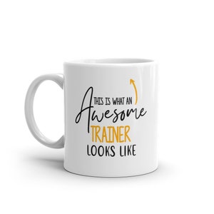 May include: White ceramic coffee mug with a black and gold design. The mug says "This is what an awesome trainer looks like" with an arrow pointing upwards.