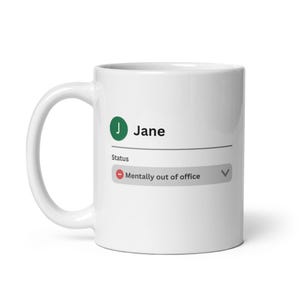 May include: White ceramic mug with the text "Jane" and a green circle with the letter "J". Below the name is the status "Mentally out of office". The mug has a curved handle.