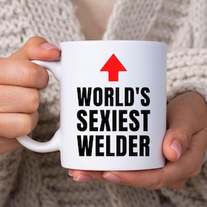 May include: A white ceramic mug featuring a red upward-pointing arrow graphic above the black text that reads WORLD'S SEXIEST WELDER. The mug has a standard handle on the left side and a smooth, cylindrical body with a clean, minimalist design.
