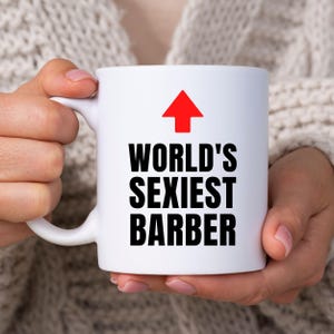 Może przedstawiać: Biały ceramiczny kubek z czerwoną strzałką skierowaną w górę nad czarnym napisem WORLD'S SEXIEST BARBER. Kubek posiada standardowe ucho po lewej stronie oraz gładki, cylindryczny korpus o prostym i przejrzystym wyglądzie zewnętrznym.