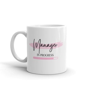 Op de afbeelding: Witte keramische mok met een roze penseelstreepontwerp en de tekst "Manager in progress" met een roze voortgangsbalk.