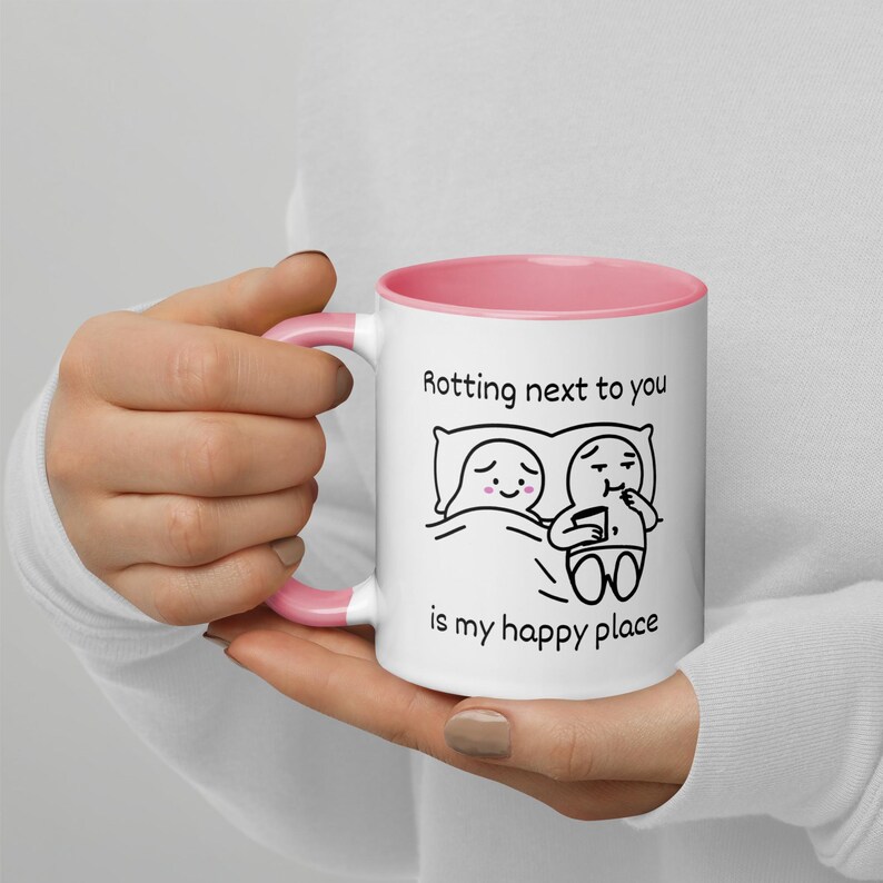 May include: White ceramic mug with a pink interior and handle. The mug features a black and white cartoon illustration of two figures in bed, with the text "Rotting next to you is my happy place."