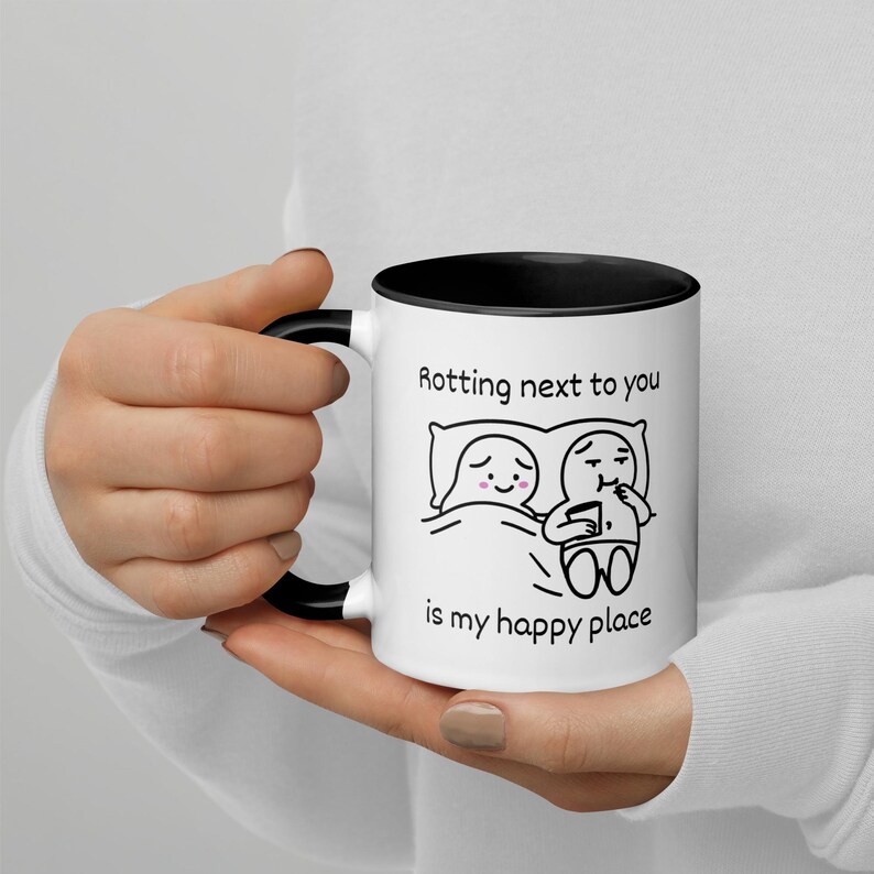 May include: White ceramic mug with a black interior and handle. The mug features a black and white cartoon illustration of two figures in bed with the text "Rotting next to you is my happy place."