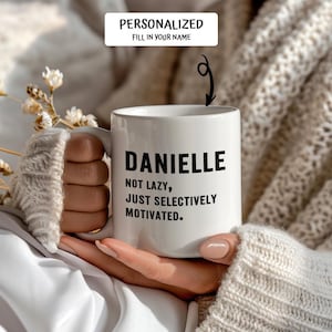 May include: White ceramic mug with the name "DANIELLE" in black, along with the text "NOT LAZY, JUST SELECTIVELY MOTIVATED." The mug is held by a person. The top of the mug has the text "PERSONALIZED FILL IN YOUR NAME".
