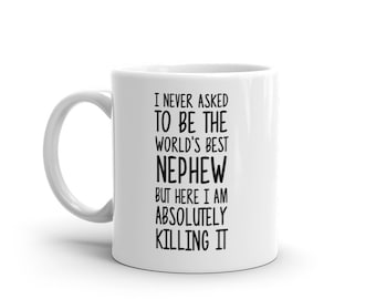 May include: White ceramic mug with a curved handle. The mug features black text that reads: "I NEVER ASKED TO BE THE WORLD'S BEST NEPHEW BUT HERE I AM ABSOLUTELY KILLING IT."