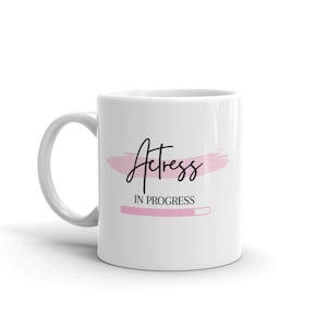 Op de afbeelding: Witte keramische mok met de woorden "Actress In Progress" in zwart script over een roze penseelstreek. Onder de tekst staat een roze voortgangsbalk. De mok heeft een gebogen handvat.
