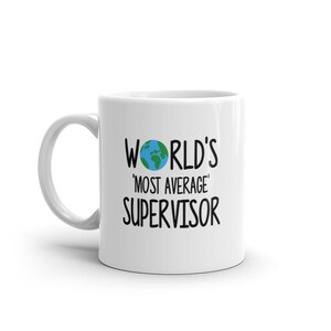 Könnte beinhalten: Weiße Keramiktasse mit einer schwarzen Grafik, auf der "World's 'Most Average' Supervisor" steht, mit einem kleinen grünen und blauen Globus über dem Text.