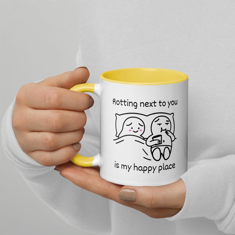 May include: White ceramic mug with a yellow interior and handle. The mug features a black and white cartoon illustration of two figures in bed, with the text "Rotting next to you is my happy place."