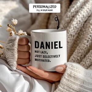 May include: White ceramic mug with the name "DANIEL" in bold black letters. Below the name, the text reads "NOT LAZY, JUST SELECTIVELY MOTIVATED." The mug is held by a person wearing a white sweater.