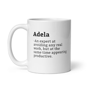 Peut inclure: Une tasse à café en céramique blanche avec un motif de texte noir qui dit "Adela -Une experte pour éviter tout travail réel, mais en même temps paraître productive."