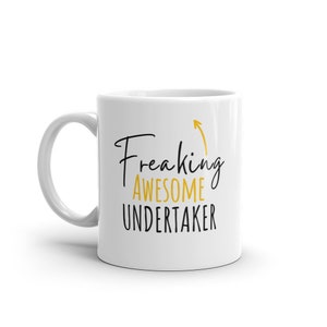 Puede incluir: Taza de café de cerámica blanca con un diseño en negro y dorado. La taza tiene un asa negra y el texto "Freaking Awesome Undertaker" con una flecha apuntando hacia arriba.