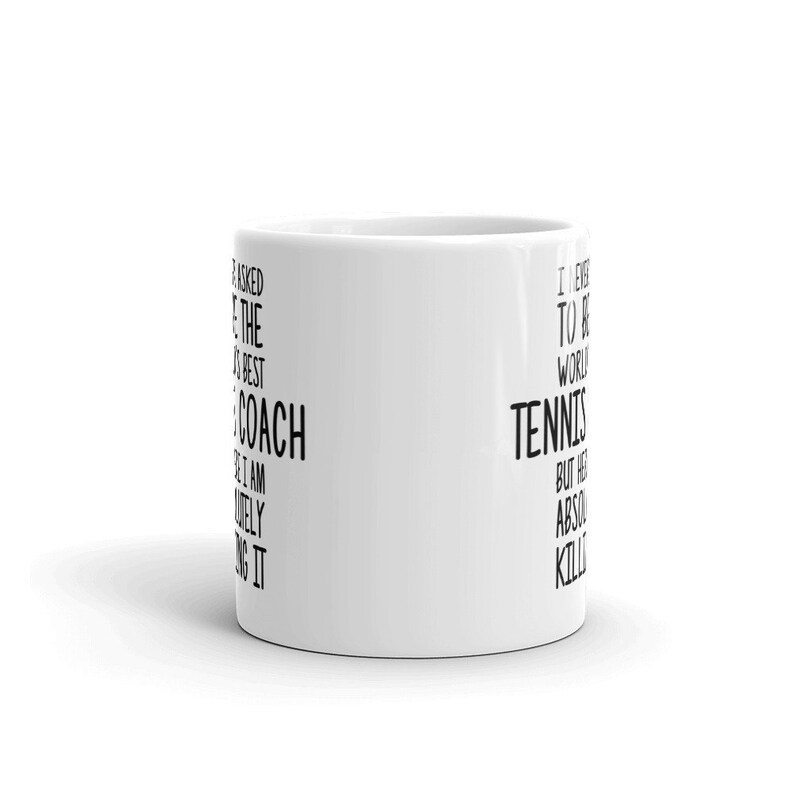 K&ouml;nnte beinhalten: Eine wei&szlig;e Keramik-Kaffeetasse mit schwarzem Text, der auf der einen Seite "Ich habe nie darum gebeten, der beste Trainer der Welt zu sein, aber ich bin absolut dabei." und auf der anderen Seite "Ich habe nie darum gebeten, der beste Tennisspieler der Welt zu sein, aber ich bringe es absolut fertig." lautet.