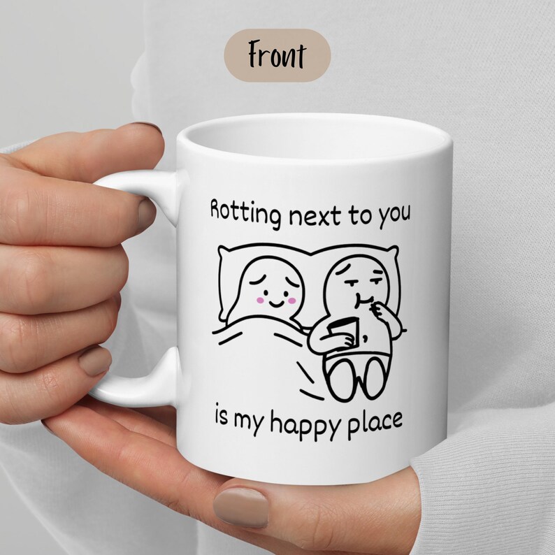 May include: White ceramic mug with a black line drawing of two cartoon figures in bed, with the text "Rotting next to you is my happy place."