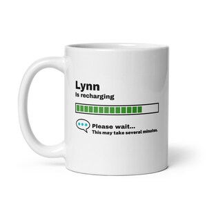 Op de afbeelding: Witte keramische mok met een zwarte en groene voortgangsbalk met de tekst "Lynn is aan het opladen". De voortgangsbalk is bijna vol. Onder de voortgangsbalk bevindt zich een spraakballon met de tekst "Wacht even... Dit kan enkele minuten duren."