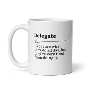 May include: White ceramic mug with the word "Delegate" in black, along with a definition. The definition reads: "- Not sure what they do all day, but they're very tired from doing it."