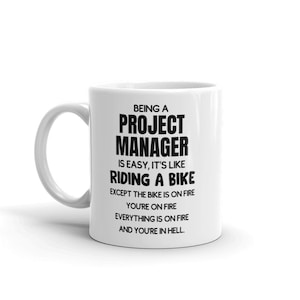 May include: White ceramic coffee mug with black text that reads "Being a Project Manager is easy, it's like riding a bike except the bike is on fire, you're on fire, everything is on fire and you're in hell."