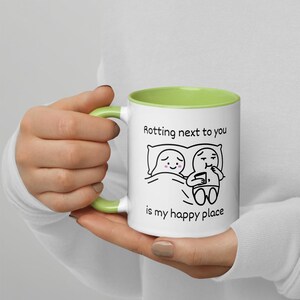 May include: White ceramic mug with a green interior and handle. The mug features a black and white cartoon illustration of two figures in bed with the text "Rotting next to you is my happy place."