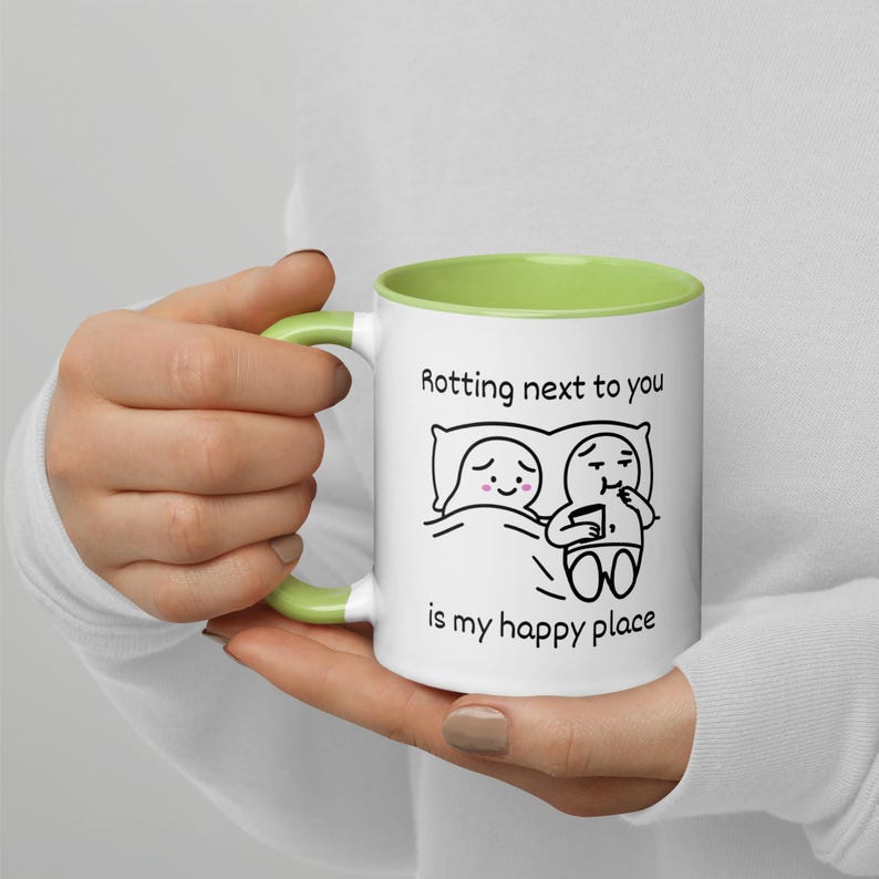 May include: White ceramic mug with a green interior and handle. The mug features a black and white cartoon illustration of two figures in bed with the text "Rotting next to you is my happy place."