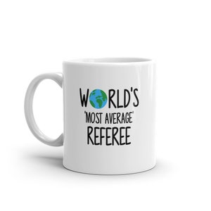Könnte beinhalten: Weiße Keramiktasse mit einer schwarzen Grafik, auf der "WORLD'S 'MOST AVERAGE' REFEREE" steht, mit einem kleinen Bild eines blauen und grünen Globus.