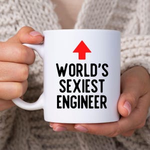 Puede incluir: Una taza de cerámica blanca con un gráfico de una flecha roja que apunta hacia arriba sobre el texto negro que dice WORLD'S SEXIEST ENGINEER. La taza tiene un acabado liso, un asa curva en el lado izquierdo y una forma cilíndrica estándar.