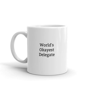 May include: White ceramic coffee mug with black text that reads "World's Okayest Delegate".
