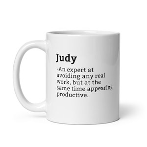 Op de afbeelding: Witte keramische koffiemok met een zwart ontwerp met de tekst "Judy - Een expert in het vermijden van echt werk, maar tegelijkertijd productief lijken."