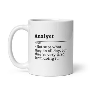 May include: A white ceramic mug with the word "Analyst" printed in black, along with a humorous definition. The definition reads: "- Not sure what they do all day, but they're very tired from doing it."