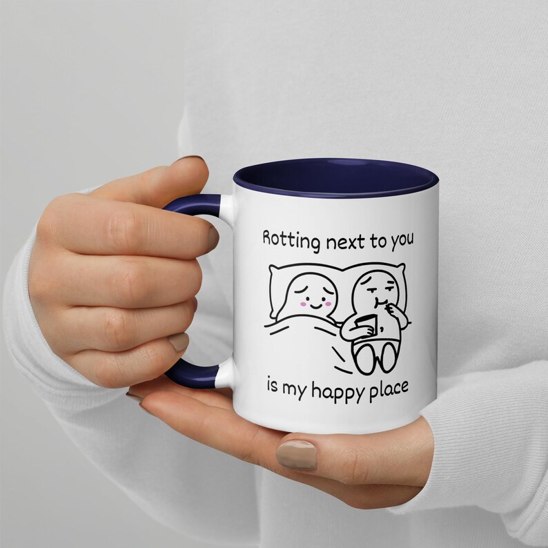 May include: White ceramic mug with a navy blue interior and handle. The mug features a black and white cartoon illustration of two figures in bed with the text "Rotting next to you is my happy place."