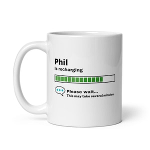 Op de afbeelding: Witte keramische mok met een zwarte en groene voortgangsbalk die "Phil is aan het opladen... Even geduld... Dit kan enkele minuten duren." aangeeft.