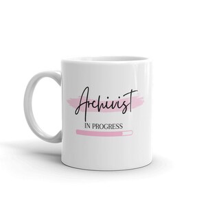 Könnte beinhalten: Weiße Keramiktasse mit einem rosafarbenen Pinselstrich-Design und dem Text "Archivist in progress" mit einer rosafarbenen Fortschrittsanzeige.