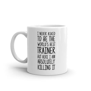 May include: White ceramic coffee mug with black text that reads "I never asked to be the world's best trainer but here I am absolutely killing it."