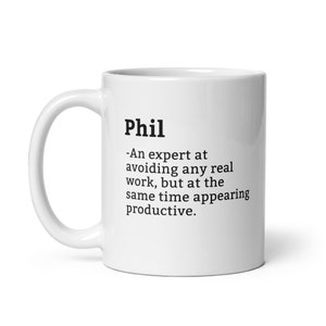 Może przedstawiać: Biały ceramiczny kubek z czarnym napisem "Phil". Poniżej humorystyczna definicja: "-Ekspert w unikaniu prawdziwej pracy, ale jednocześnie sprawiający wrażenie produktywnego."