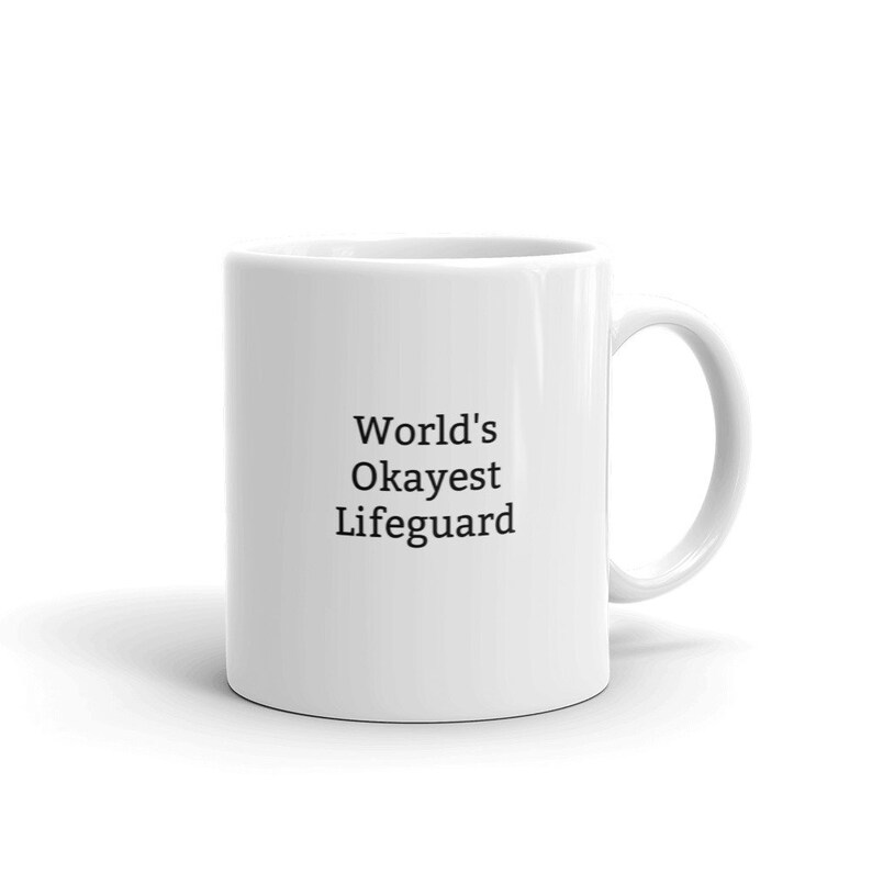 May include: White ceramic coffee mug with black text that reads "World's Okayest Lifeguard".