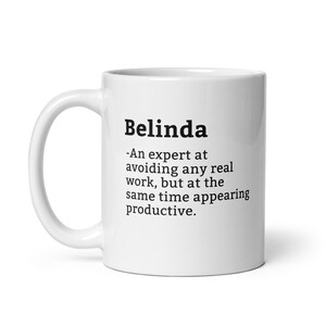 Könnte beinhalten: Weiße Keramik-Kaffeetasse mit einem schwarzen Textdesign, das "Belinda - Eine Expertin darin, jede wirkliche Arbeit zu vermeiden, aber gleichzeitig produktiv zu erscheinen." lautet.