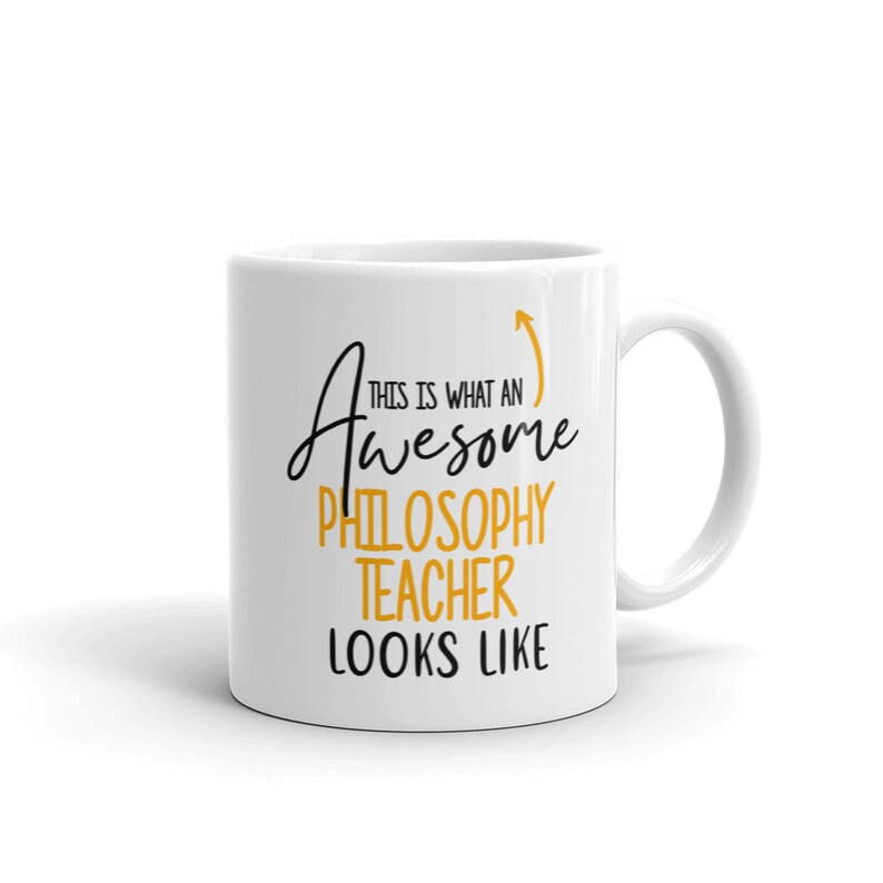 以下が含まれることがあります： 白のセラミックマグカップ。黒と金のテキストデザインで、「This is what an Awesome Philosophy Teacher Looks Like」と上向きの矢印が描かれています。 マグカップには湾曲したハンドルが付いています。