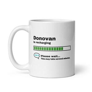 May include: White ceramic mug with a black and green progress bar that reads "Donovan is recharging". The progress bar is almost full. Below the progress bar is a speech bubble with three blue dots and the text "Please wait... This may take several minutes."