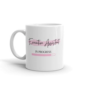 Könnte beinhalten: Weiße Keramiktasse mit den Worten "Executive Assistant" in schwarzer Schrift und "IN PROGRESS" darunter. Auf der Tasse befindet sich auch eine rosa Fortschrittsanzeige. Die Tasse hat einen gebogenen Henkel.