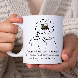 May include: White ceramic mug featuring a cartoon illustration of two stick figures and a thought bubble with a train. The text on the mug reads, "Dave might look like he's listening but he's actually thinking about trains."