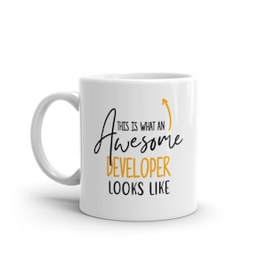 Peut inclure: Une tasse à café en céramique blanche avec un motif noir et or. Le texte sur la tasse est "This is what an awesome developer looks like".