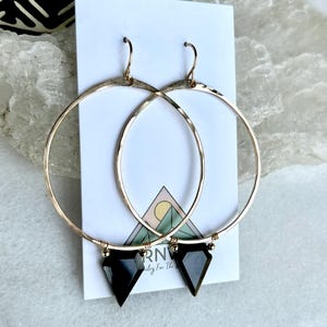 May include: Gold-tone hoop earrings with black geometric pendants. The earrings are displayed on a white card with a logo and a mountain graphic. The hoops have a hammered texture and are suspended from gold-colored ear wires.