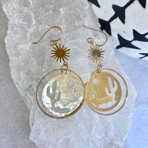 May include: Gold tone earrings with a desert scene cutout. The earrings feature a circle with a cactus, mountains, and a moon. The top of the earrings have a starburst design.