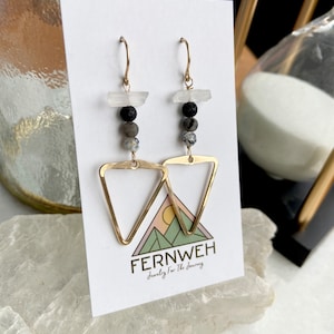 May include: Gold-tone triangle earrings with a layered design. The earrings feature clear, black, and gray beads, and a triangular gold-tone pendant. The earrings are displayed on a white card with the brand name "FERNWEH" and a mountain graphic.
