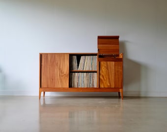 May include: A mid-century modern wooden record cabinet with a sliding door and a flip-top compartment. The cabinet is made of warm-toned wood and holds numerous vinyl records. The cabinet is set against a white wall.