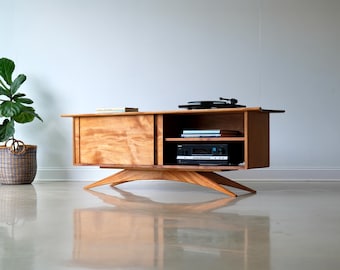 May include: A mid-century modern wooden media console with a sliding door and open shelving. The console features a turntable on top and a stereo receiver on the shelf. The console is supported by a unique, angled base.