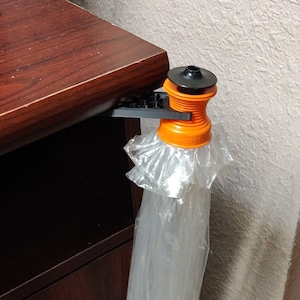 Peut inclure: Un porte-sac en plastique noir et orange fixé au bord d'un bureau en bois. Le porte-sac est muni d'un sac en plastique transparent qui pend.